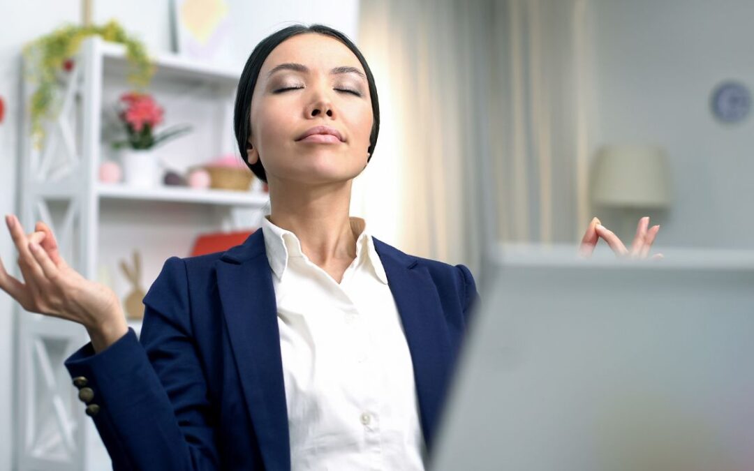 Stress and Anxiety in Business: a photo of a woman in her office, at her desk closing her eyes and meditating.